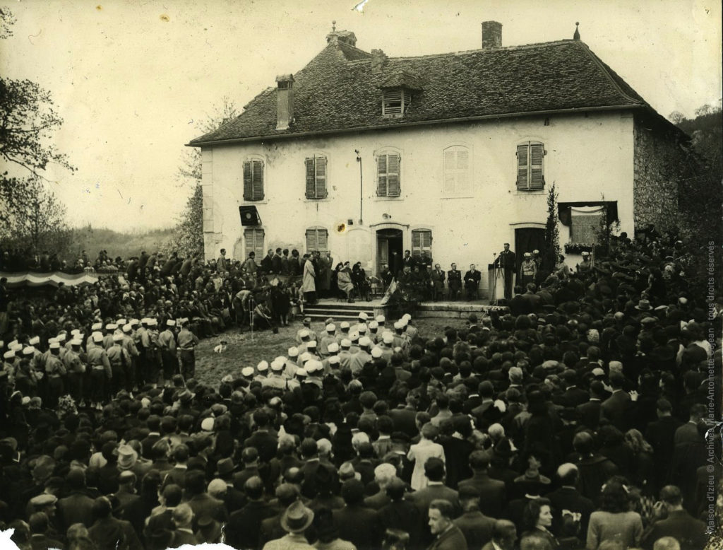 © Archives Marie-Antoinette Cojean - CAG Sur la terrasse, à gauche de la plaque, les officiels : le ministre des Victimes de guerre, le chef de cabinet représentant le ministre de la Population, le préfet, le président du Conseil général, les quatre députés de l’Ain, le secrétaire général du ministère des Victimes de guerre, le sous-préfet de Belley. Puis viennent les membres du Comité d’honneur, les invités officiels et les membres du Comité organisateur. « Au pied de la terrasse côté plaque, un groupe d’éclaireurs de France …/… la presse, la radiodiffusion et les actualités, les délégations de Déportés, Prisonniers, anciens du Maquis. ». La musique est assurée par la fanfare des Avenières augmentée de quelques membres de la fanfare des pompiers de Brégnier-Cordon. À droite de la plaque : les enfants des écoles de Brégnier-Cordon, d’Izieu et de Belley, les municipalités d’Izieu et Brégnier-Cordon et les invités. Devant la maison, face aux marches, le 1er bataillon du 1er Régiment de Tirailleurs Marocains de Bourg-en-Bresse rend les honneurs. La cérémonie est sonorisée et filmée, le cameraman vérifie sa prise de vue en bas des marches à gauche. Le site est pavoisé aux couleurs de la toute nouvelle République, une longue bande de tissu tricolore se déploie le long de la rambarde de la terrasse, de même que sur le pupitre des orateurs. « Une sorte de chapelle de verdure » a été créée autour de la plaque, des tissus tricolores et noirs parachèvent cette scénographie.